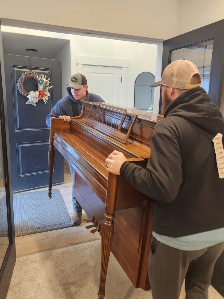 Two men from All Smile Moving carry a piano into a house, demonstrating teamwork and careful handling of the instrument.