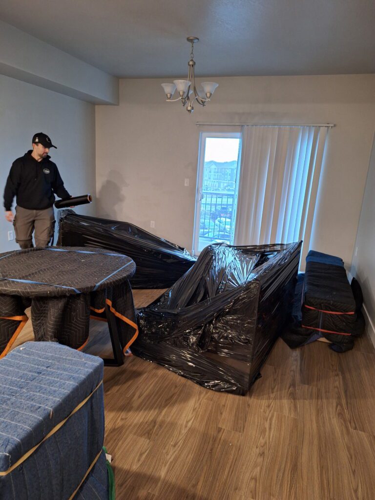 A person wraps furniture with black plastic in a room with wooden floors. The table and chairs are covered with moving blankets. A window with blinds lets in natural light.