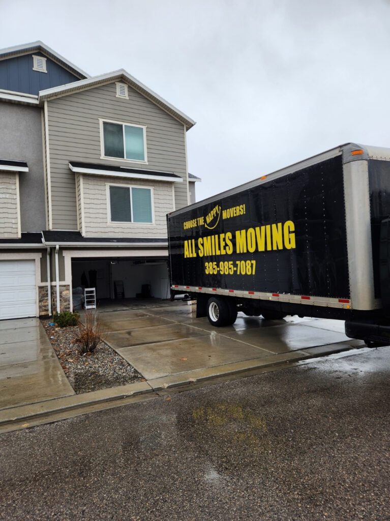 A moving truck labeled "All Smiles Moving" is parked in the driveway of a gray townhouse on a rainy day. The garage door is open, showing moving items.