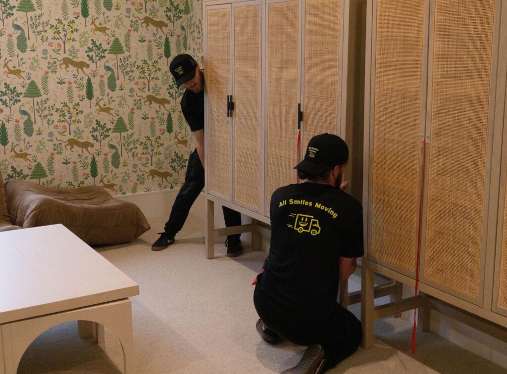 Two movers in branded shirts are relocating a cabinet in a child’s room with dinosaur wallpaper. They appear focused and carefully coordinate the move.