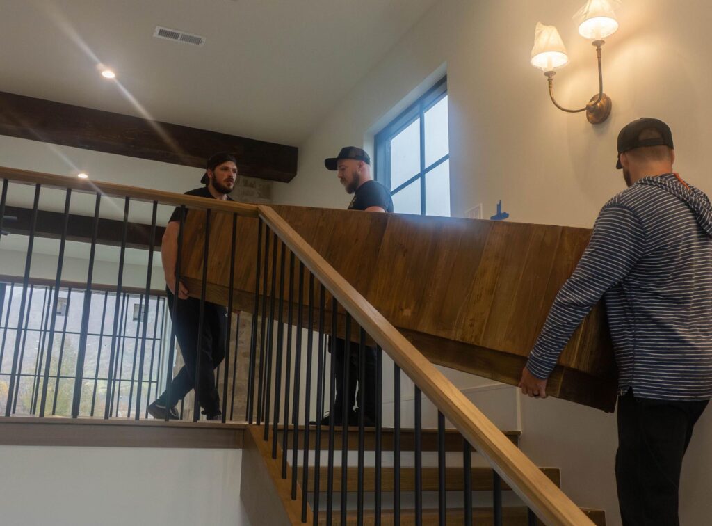 Three men carry a large wooden table up a staircase in a modern home. They work together, showing concentration and teamwork in a well-lit space.
