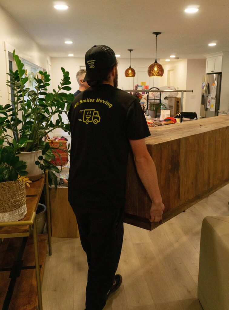 A mover in a black "All Smiles Moving" shirt carries a large wooden table in a warmly lit living room with plants and modern decor.