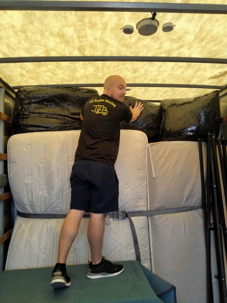 A person stands on a padded surface inside an All Smile Moving truck, securing large wrapped items and mattresses stacked against the walls.