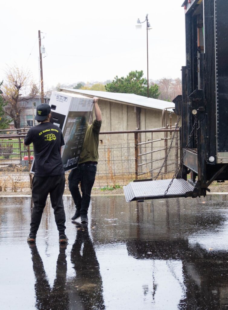 Two movers in rainy weather unload a large boxed TV from a truck using a lift gate. The reflection of the scene is visible on wet pavement.