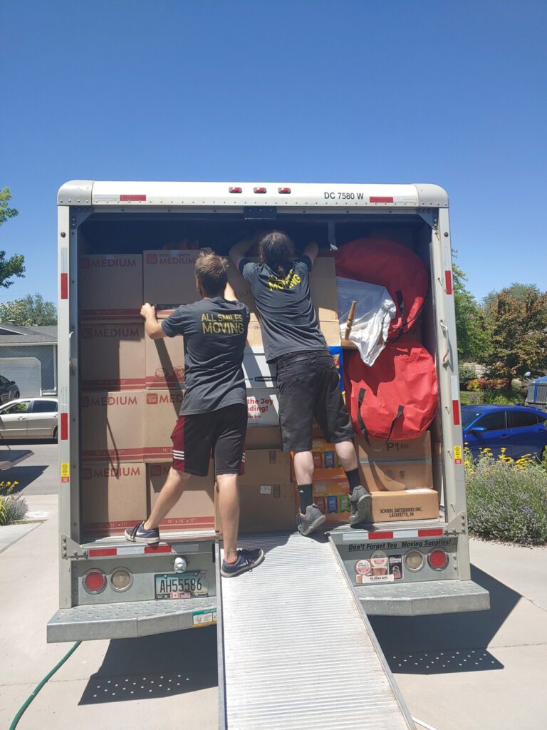 Two men unloading boxes from a moving truck labeled "All Smile Moving" on a sunny day.