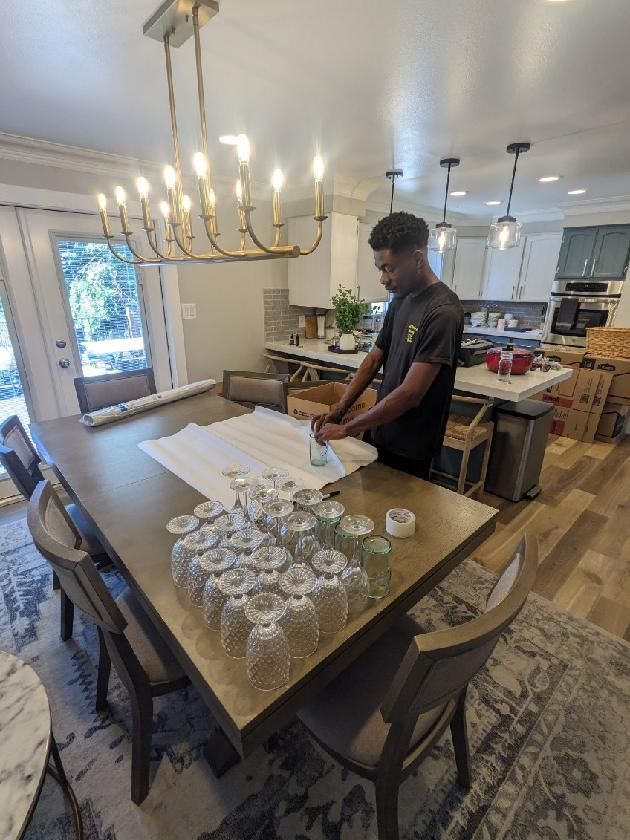 A man wraps glassware in a kitchen-dining area, with glasses and packing materials spread on the table and All Smile Moving boxes stacked in the background.