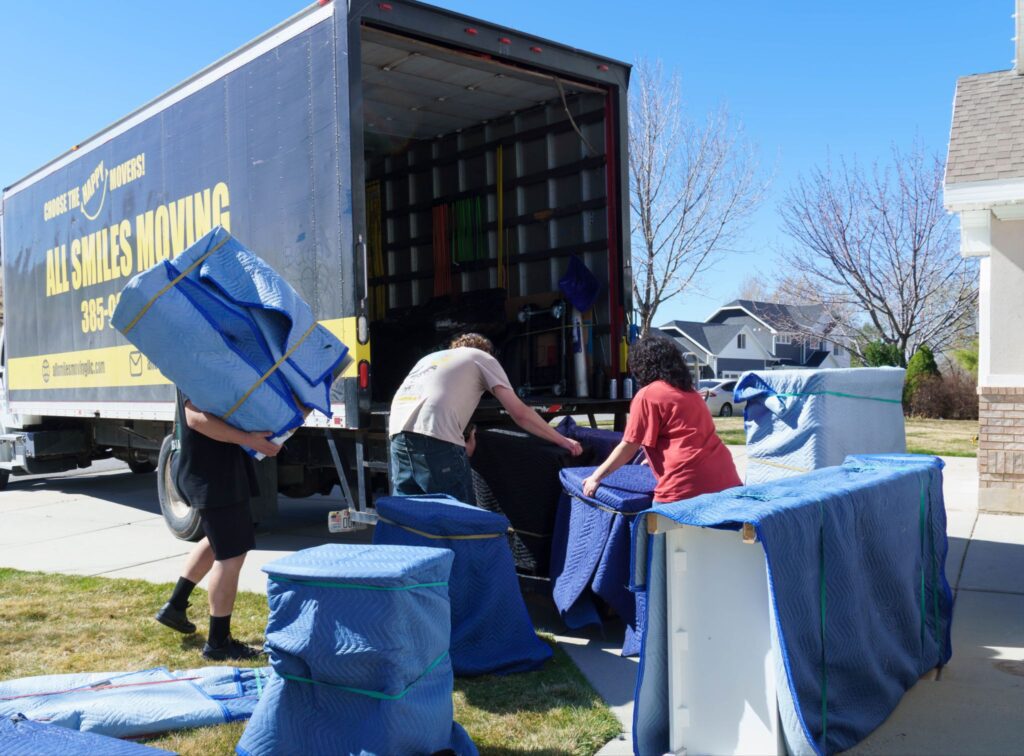 Movers loading padded furniture into the open back of an All Smiles Moving truck outside a home.